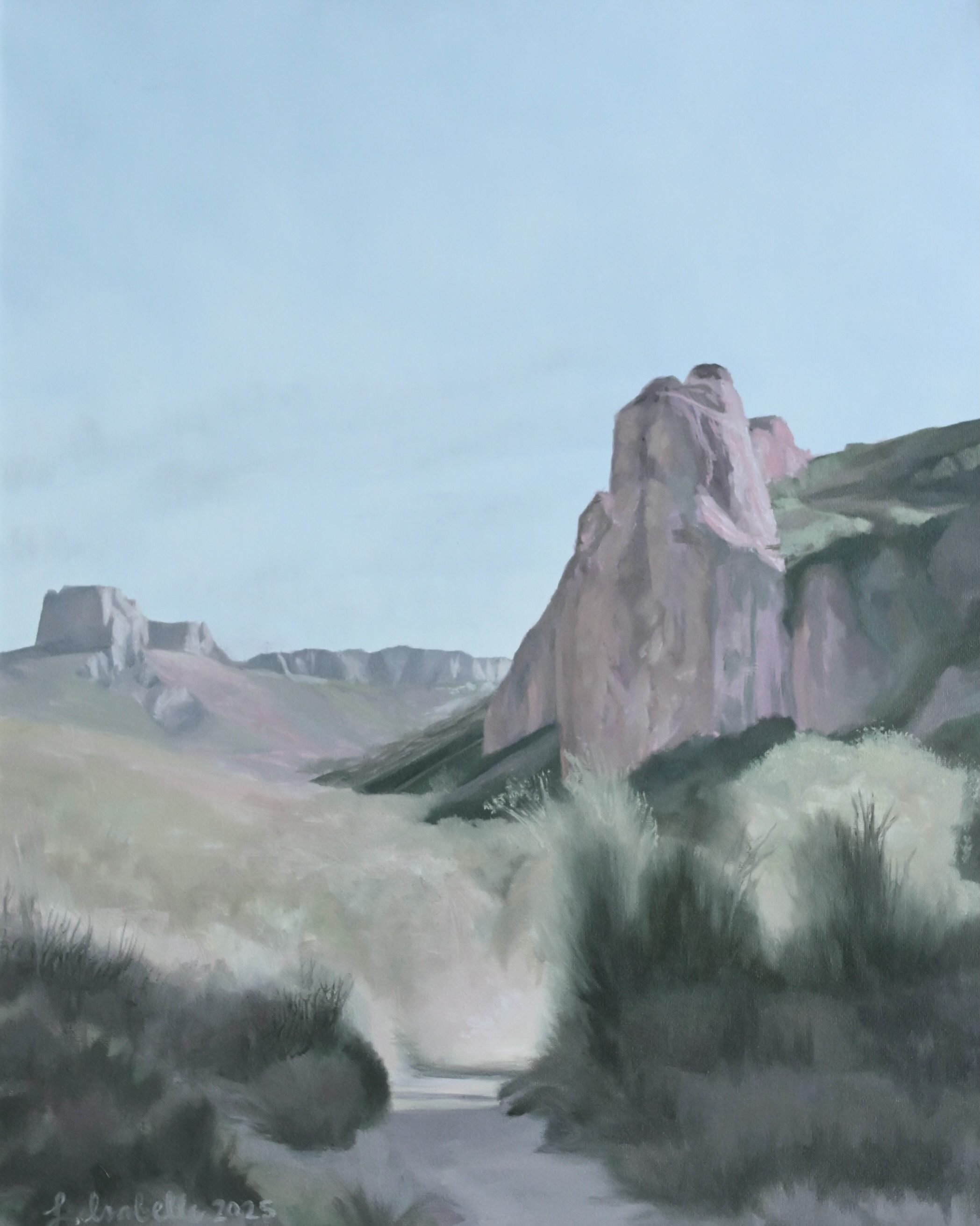 A pale desert valley with a tall pink rock tower rising on the right, flat-topped bluffs in the distance, and a sandy wash running down through grey-green scrub.